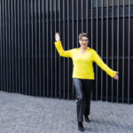 Sally Farrant in a yellow jumper and black trousers against a black gate
